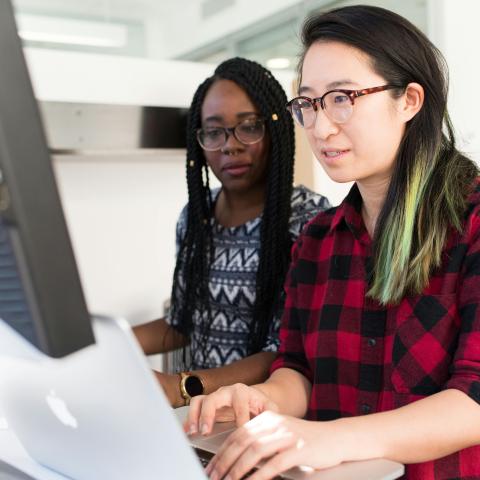 Two women working together at a desk, looking at a computer screen. One is typing on a laptop while the other observes, both focused on the task.
