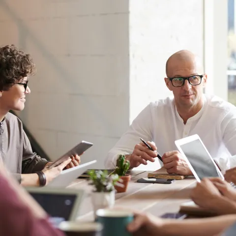 A diverse group of people engaged in a focused meeting at a bright office table. They use tablets and laptops, conveying a collaborative atmosphere.