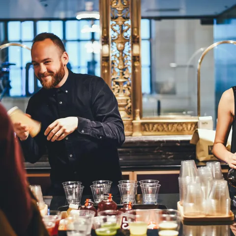 Two hospitality staff members working behind a bar — one smiling and shaking a cocktail while the other prepares a coffee — surrounded by cups, bottles, and bar equipment in a warmly lit setting.