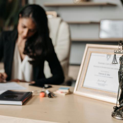 A woman in a black blazer writes at a desk with a "Lady Justice" statue and framed certificate in the foreground, conveying focus and professionalism.