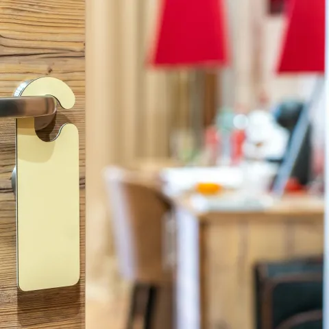 Wooden door with a metal handle and blank door hanger. Inside, a cozy room with a blurred desk, chairs, and red lamps, conveying warmth.