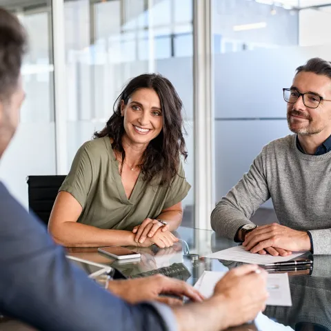 A smiling couple in a meeting with a man at a glass table in a bright office. They appear engaged and relaxed, suggesting a positive discussion.