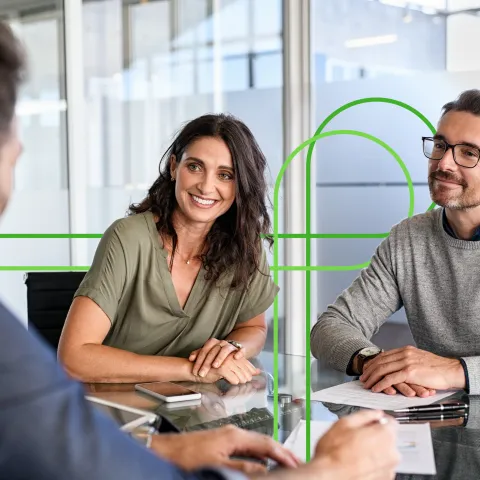 A cheerful couple sits across a table from a businessman in an office setting, engaged in a friendly conversation. The atmosphere is positive and collaborative.