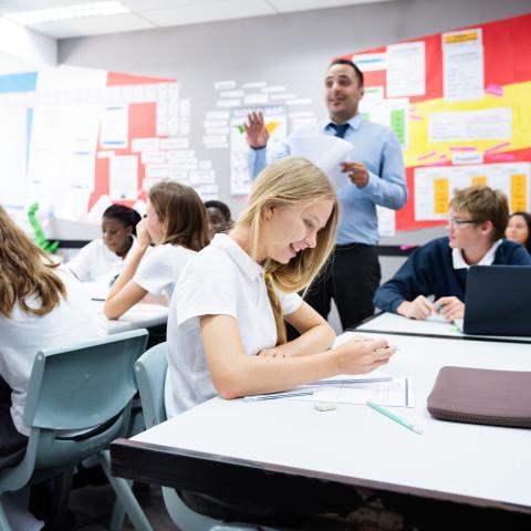 A classroom of secondary school students engaged in a lesson, with a teacher standing at the front speaking while students work at their desks using notebooks and laptops.