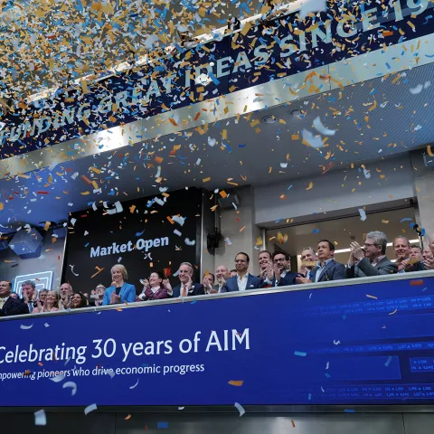 A group of people celebrate 30 years of AIM at a stock exchange. Blue confetti rains down as they stand behind a blue banner with digital displays.