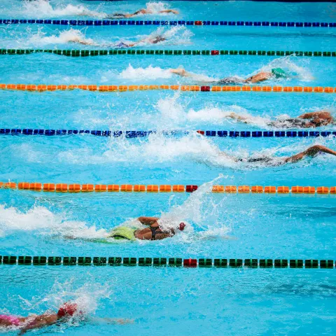 Competitive swimmers race in lanes of a pool, creating splashes as they perform freestyle strokes. The scene conveys intensity and focus.
