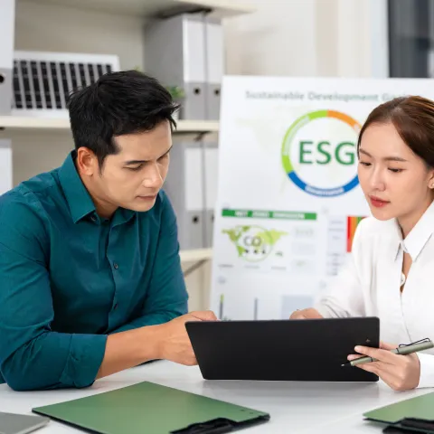 Two people in a modern office discuss sustainability. A laptop and ESG poster are visible, reflecting focus and collaboration. Shelves with plants in background.