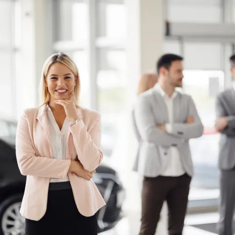 A confident woman in a pink blazer smiles in the foreground of a bright office. In the background, colleagues engage in conversation near a vehicle.