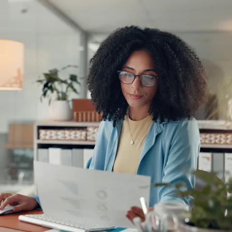 A woman with glasses and curly hair works at a desk in a modern office, focused on a document. Surroundings include plants, a lamp, and a computer.