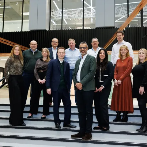 A group of team members from UHY Hacker Young and Affinia's leadership teams, both men and women, stand on indoor stairs in business attire, smiling. The background shows a modern office space with large windows.