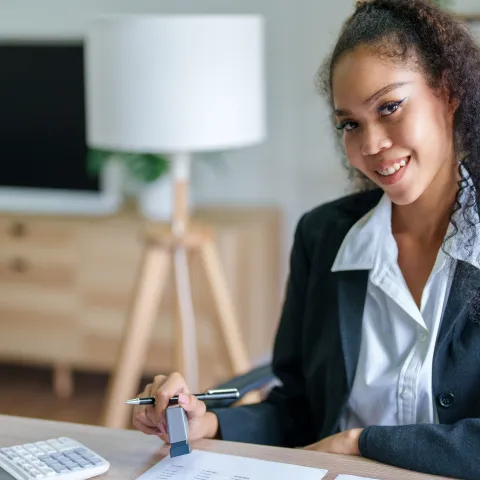 A smiling woman in a business attire sits at a desk, holding a pen. Behind her, a lamp and shelves create a professional and welcoming atmosphere.