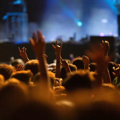 A lively concert scene with blurred crowds raising hands, enjoying vibrant music. Blue stage lights illuminate a performer in the background, creating an energetic atmosphere.