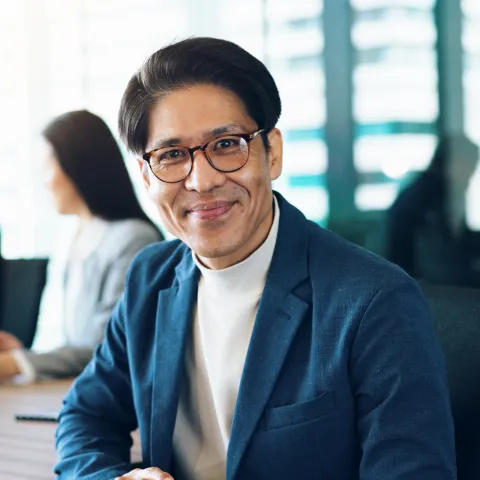 Smiling man in a blue blazer and glasses sits at a table in a bright conference room. Two colleagues are blurred in the background. Professional and welcoming tone.