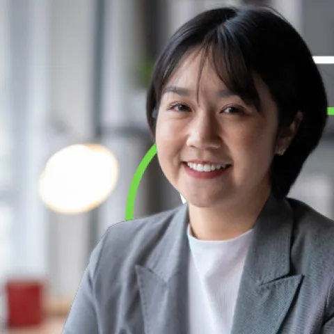 Smiling person in a gray blazer, sitting at a desk in an office. A blurred lamp and red mug are in the background, with green geometric lines overlaying.