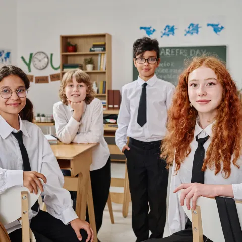 Four students in school uniforms smile confidently in a classroom. Bookshelves and a chalkboard with "careers day" add an academic atmosphere.
