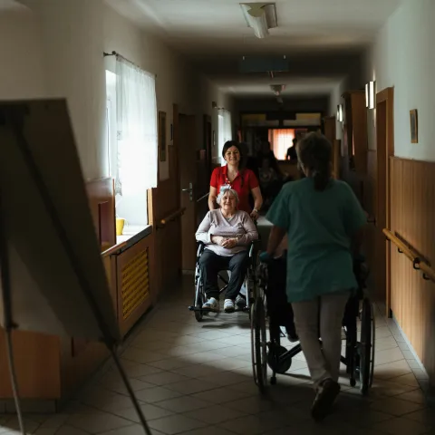 A caregiver in red pushes an elderly woman in a wheelchair down a dimly lit hallway, conveying a calm, caring atmosphere in a nursing home setting.