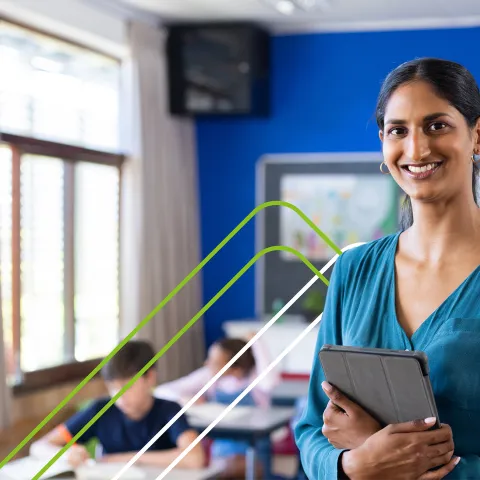 Smiling teacher in a teal blouse holds a tablet in a bright classroom with students working at desks. The atmosphere is positive and engaging.
