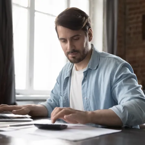 A man in a casual blue shirt works from home, focused on using a calculator and laptop. The scene is well-lit, with a brick wall in the background.