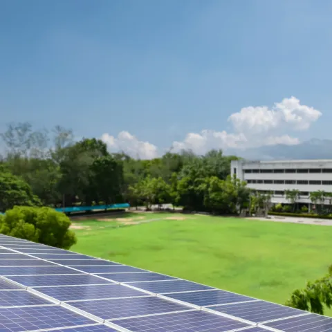 Solar panels in the foreground under a clear blue sky, with a lush green field and a distant building surrounded by trees, conveying sustainability.