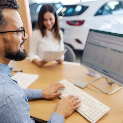 Man at a desk using a computer with a spreadsheet open, smiling woman in the background holding papers. Car dealership setting, bright and professional.
