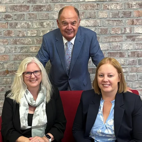 Fiona Davey, Mel Stone and Vince Edwards, partners of UHY Peninsula, smiling indoors against a brick wall. Two women sit on a red couch, while a man stands behind them. All wear business attire.