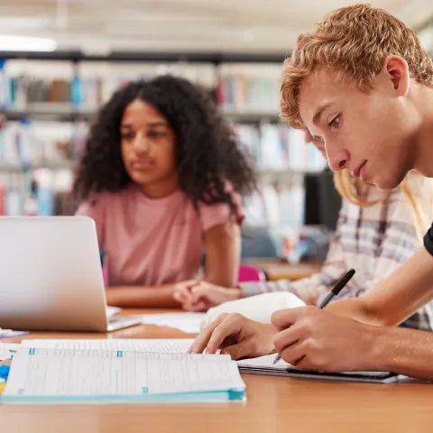 A group of focused students study at a library table. A young man takes notes, while a woman works on a laptop. Shelves of books are in the background.