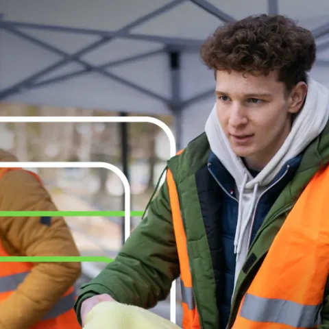 Young person in an orange safety vest and green jacket under a canopy, looking focused. Another person in a similar vest is partially visible beside them.