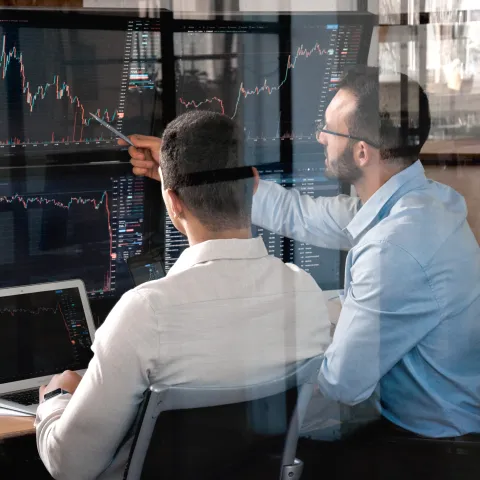Two men sit at a desk with multiple screens displaying stock market charts. One points at trends with a pen, conveying focus and collaboration.
