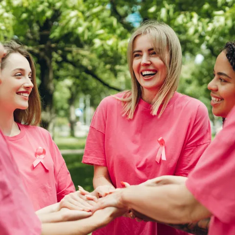 Five women in pink shirts with ribbons stand in a circle outdoors, smiling and stacking hands in solidarity, conveying unity and support.