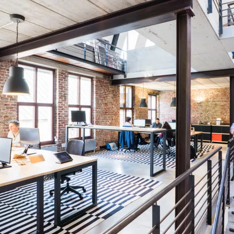 Modern open-plan office with exposed brick walls, steel beams and a mezzanine level. Several people work at desks with computer monitors, while a blurred figure walks up a concrete staircase on the right. Large windows provide natural light and black pendant lamps hang above the workspace. Black-and-white striped rugs define the seating areas.
