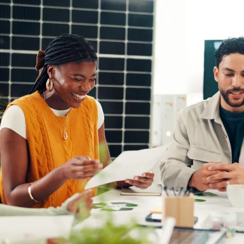 A diverse team of professionals smiling and reviewing documents together during an environmental presentation in a modern office with a solar panel background.