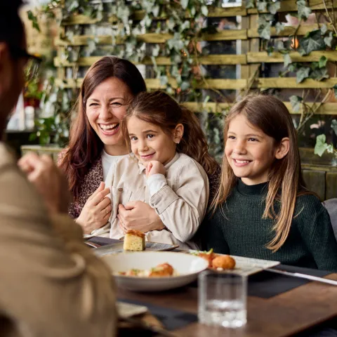 A smiling mother and her two young daughters sitting at a restaurant table, looking at a man in the foreground.