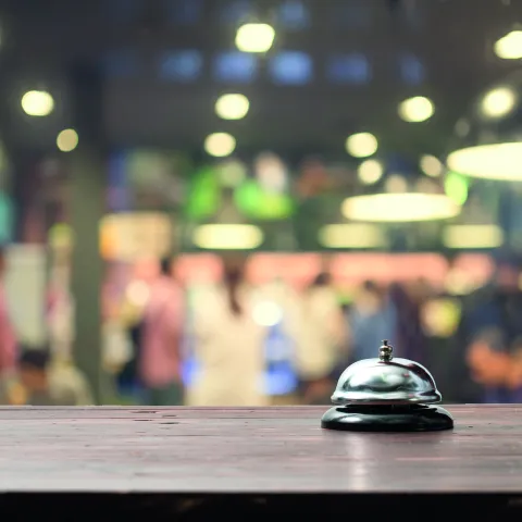 Silver service bell sitting on a wooden countertop with a blurred hospitality or reception area in the background, illuminated by warm overhead lights.