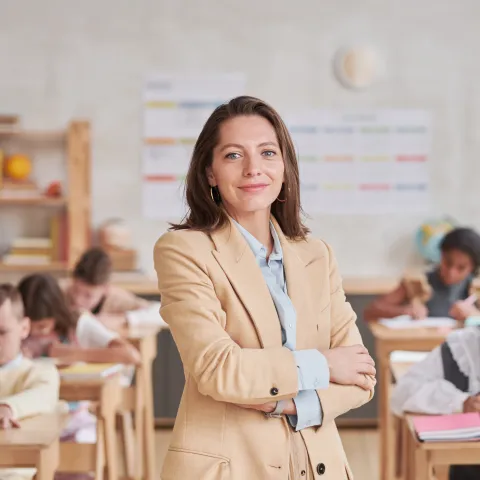 A woman wearing a beige blazer stands with arms crossed, smiling at the camera in a classroom, with children working at desks and educational posters, a globe, and a bookshelf visible in the background.