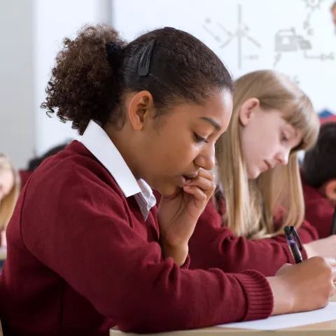A schoolgirl in a maroon jumper writes at her desk with a concentrated expression, resting her chin on her hand, while classmates in matching uniforms work alongside her and a teacher writes on a whiteboard in the background.