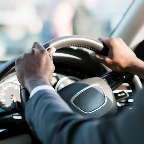 Close-up of a man in a suit gripping the leather-trimmed steering wheel of a car with both hands, with the dashboard, speedometer, and blurred street scene visible in the background.