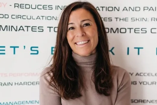 A woman with long brown hair, wearing a taupe turtleneck sweater and black trousers, sits smiling in front of a white wall covered with wellness-related text. Phrases on the wall include words like "MIND," "BODY," "PAIN MANAGEMENT," and "AESTHETIC," in various colours and fonts, highlighting the benefits of cryotherapy and wellness treatments.
