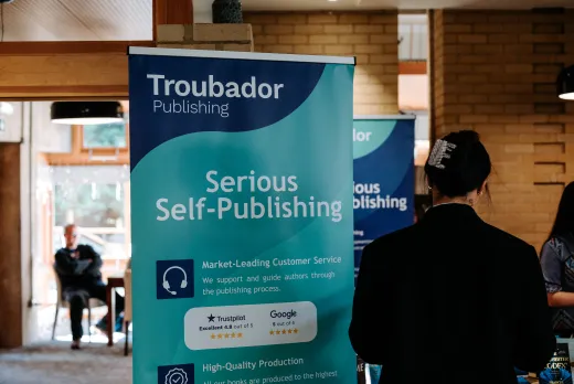 Banner for Troubador Publishing promotes 'Serious Self-Publishing' in an indoor setting. A woman stands nearby, suggesting an event or seminar atmosphere.
