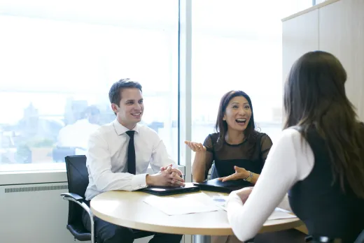 Three people in business attire are engaged in a lively discussion around a round table in a bright office. Two face the camera, smiling and gesturing animatedly.