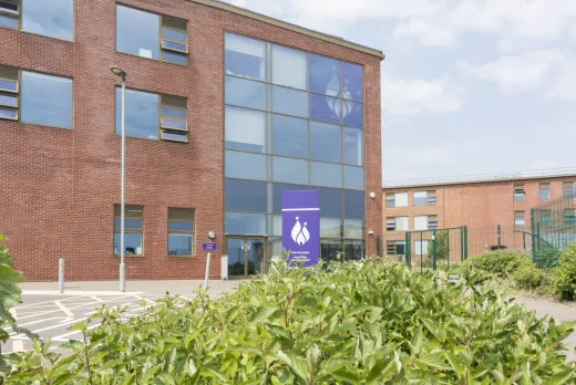 Exterior view of a modern brick school building with large glass windows and a purple sign near the entrance, surrounded by greenery.
