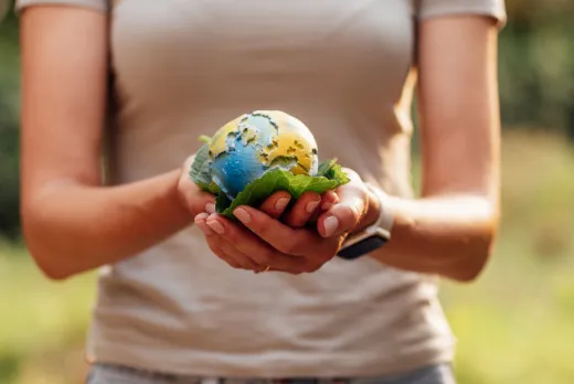 A person holds a small globe resting on green leaves, symbolizing environmental care. The background is a blurred, lush natural setting, creating a peaceful tone.