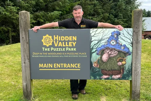 A man wearing a black polo shirt with the Hidden Valley logo stands behind a large wooden entrance sign, resting his arms along the top edge. The sign reads "Hidden Valley, The Puzzle Park – Deep in the woodland is a puzzling place where families and friends will find mysteries to chase – Main Entrance," and features an illustrated forest troll wearing a tall blue mushroom-cap hat covered in eyes. The sign stands on a grassy area with tall evergreen trees in the background.