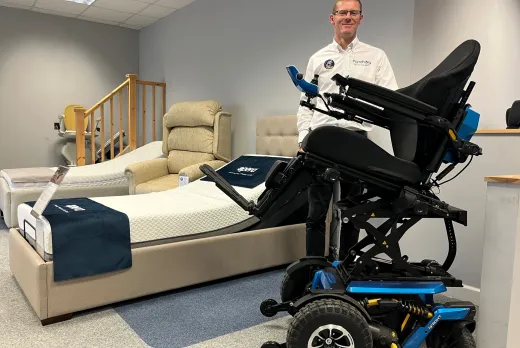 A man in a white shirt stands in a mobility showroom next to an advanced power wheelchair with large all-terrain wheels and a raised, tilted seat. Behind him, the showroom displays adjustable beds, a stairlift and mobility armchairs.