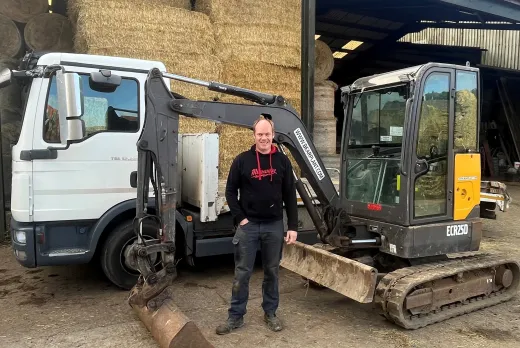A person standing in front of a flatbed truck and a compact excavator inside a farmyard. Large stacks of hay bales are stored in a covered barn behind the machinery, with agricultural equipment and farm buildings visible throughout the scene.